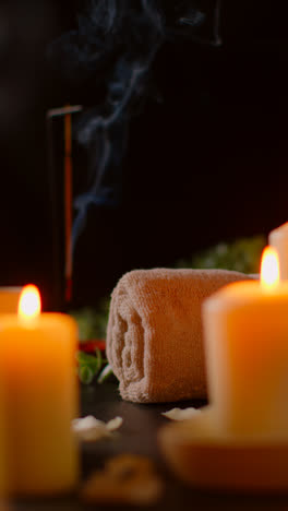 Vertical-Video-Shot-Of-Person-Picking-Up-Towel-With-Lit-Candles-With-Scattered-Petals-Incense-Stick-Against-Dark-Background-As-Part-Of-Relaxing-Spa-Day-Decor